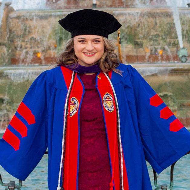 Aly Weitkamp posing in front of a fountain and wearing blue and red commencement regalia after graduating from law school at DePaul University in Illinois.