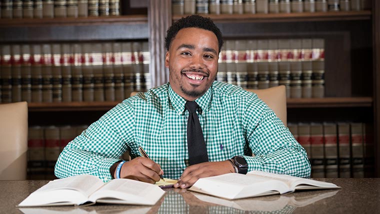 A pre-law student looks up to smile while taking notes from two big textbooks.