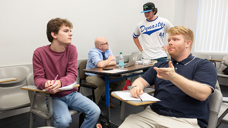 Two students seated at their desks have a discussion during their graduate history education class. In the background, Dr. Travis Seay gives feedback to a student on his presentation.