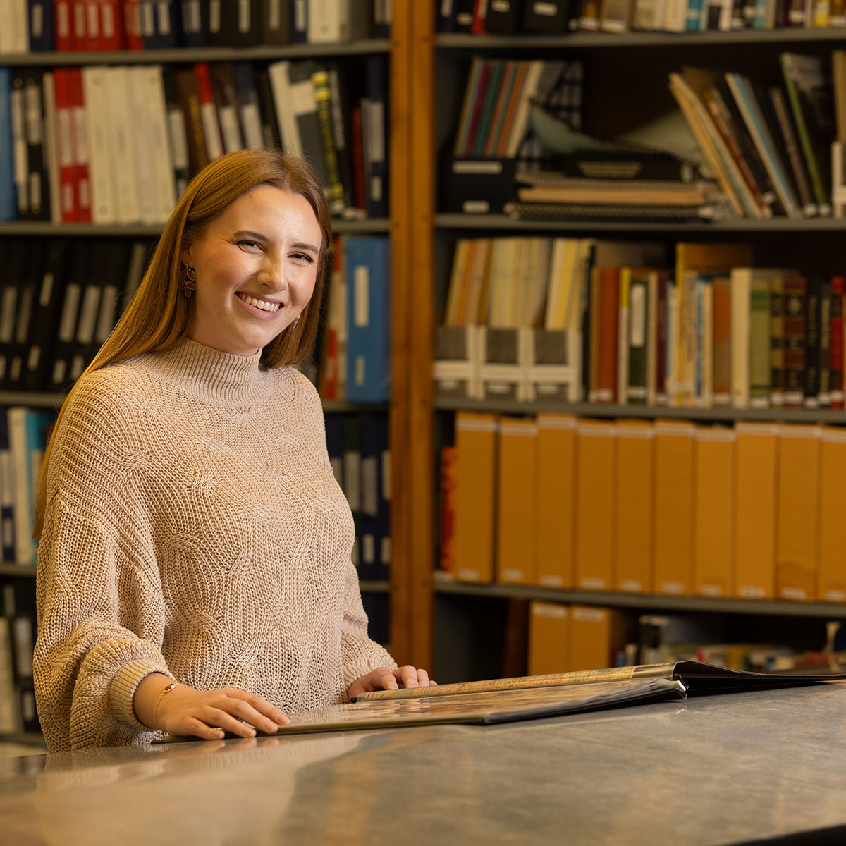 Susan Hardy smiles at the camera while opening a book in front of the collections at the Greene County Archives.